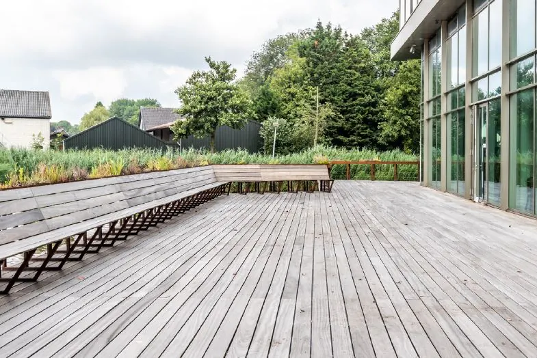 Houten terras met lange houten bank bij een modern gebouw aan het John M. Keynesplein, omringd door groen.